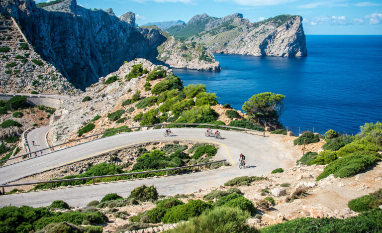 Ciclistas sobem ladeira pavimentada no litoral rochoso de Mallorca.