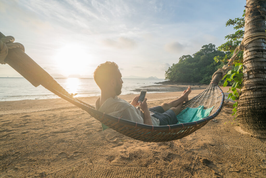 Homem de short relaxa em rede na praia enquanto navega no celular.
