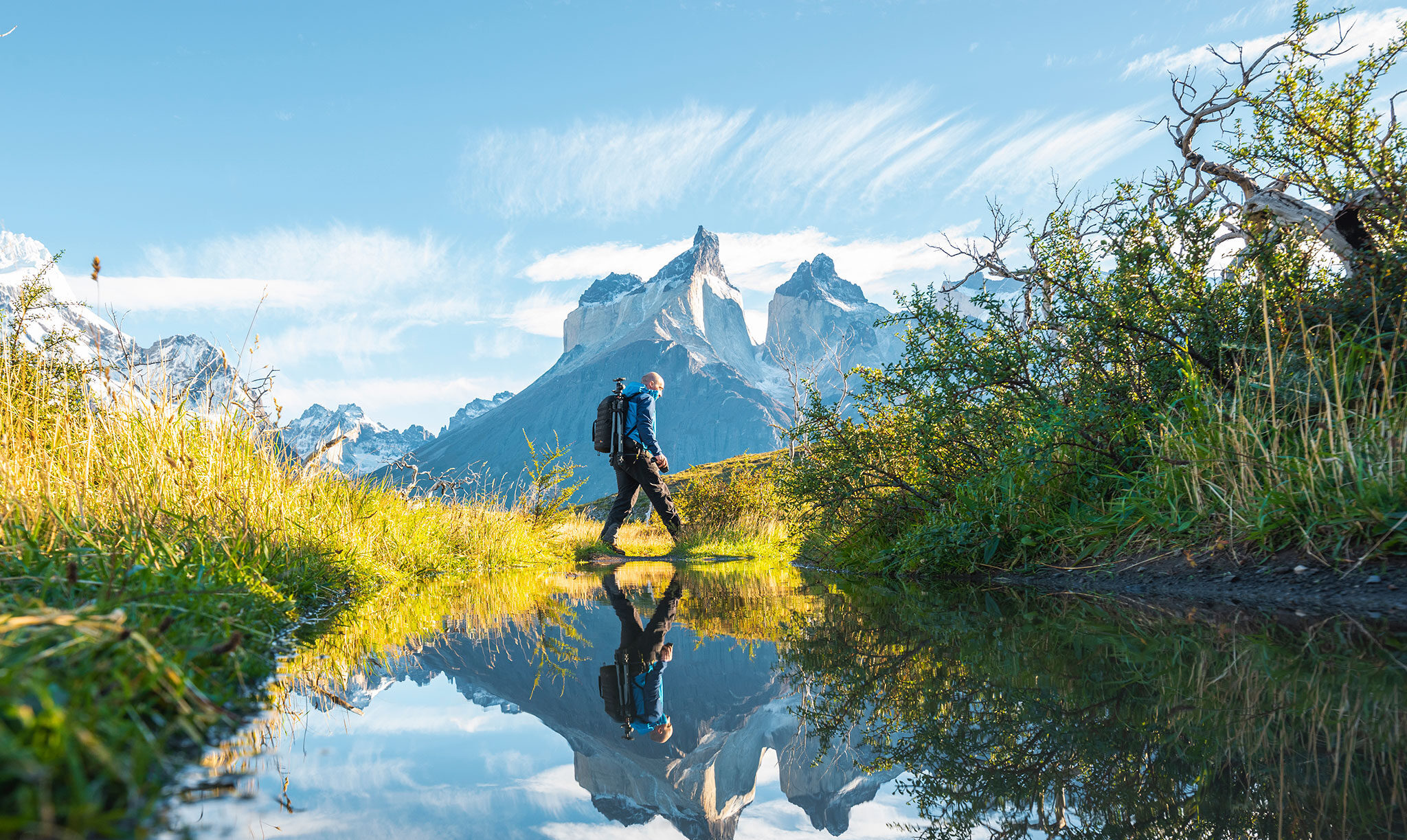 Viajante com equipamento fotográfico atravessa um açude diante de montanhas com picos agudos ao fundo.